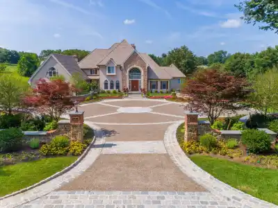 A large, elegant house with a stone facade stands at the end of a circular driveway. Surrounded by expertly crafted gardens and neatly trimmed hedges, thanks to premier landscaping services Delaware County PA, it basks under the blue sky and lush greenery in this serene setting.