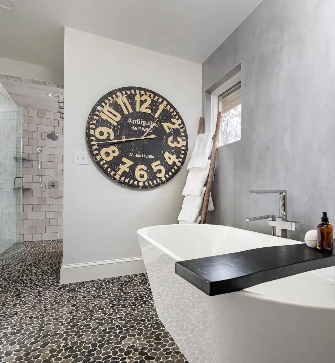 Modern bathroom with a freestanding white tub and large black and white wall clock, reminiscent of the elegance found in top landscaping services Delaware County PA offers. Pebble-style tiles cover the floor, complemented by a towel ladder rack. The shower area boasts white tiled walls and a sleek glass door.