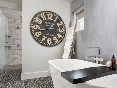 Modern bathroom with a freestanding white tub and large black and white wall clock, reminiscent of the elegance found in top landscaping services Delaware County PA offers. Pebble-style tiles cover the floor, complemented by a towel ladder rack. The shower area boasts white tiled walls and a sleek glass door.