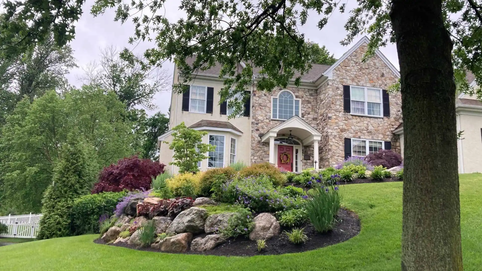 A two-story house with stone and beige siding stands amidst lush landscaping, a testament to premier landscaping services in Delaware County, PA. The front yard boasts a manicured lawn, vibrant flower beds, rocks, and trees under a cloudy sky, with a white fence on the left.