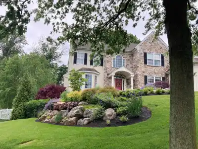 A two-story house with stone and beige siding stands amidst lush landscaping, a testament to premier landscaping services in Delaware County, PA. The front yard boasts a manicured lawn, vibrant flower beds, rocks, and trees under a cloudy sky, with a white fence on the left.