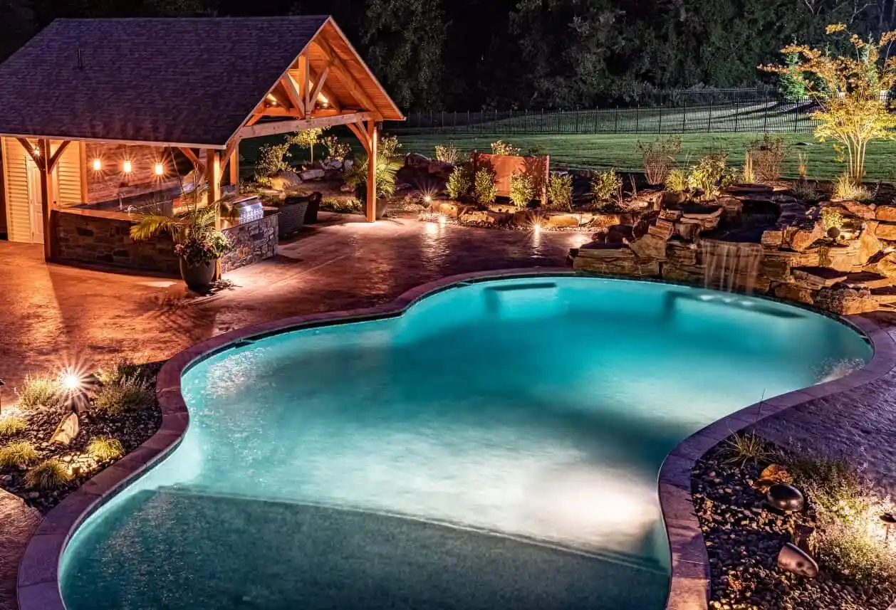 In a serene evening ambiance, an illuminated backyard scene showcases a glowing pool with a waterfall. Beside it, a rustic pavilion featuring a bar and string lights enhances outdoor living in Delaware County, PA. Landscaped gardens and trees surround the area, completing the picturesque setting.