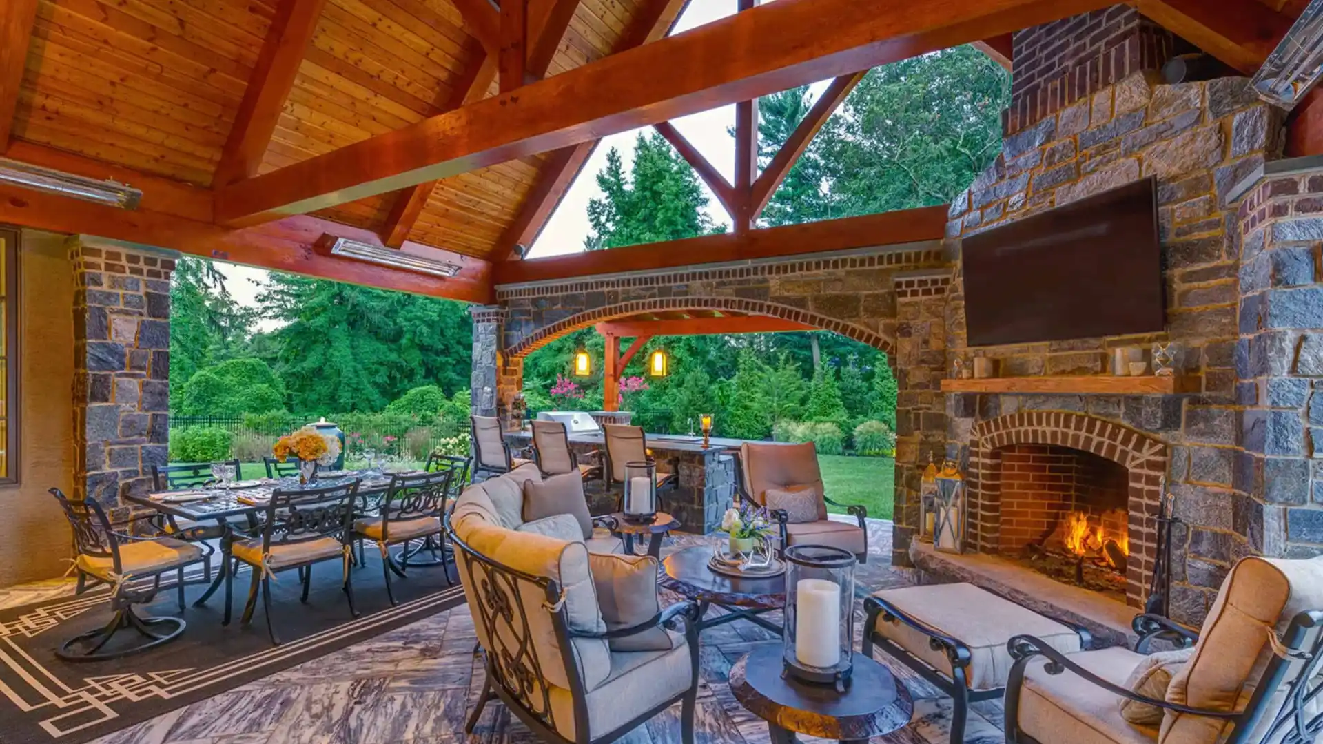 Luxurious outdoor patio with wooden ceiling and stone walls, featuring cushioned seating around a fireplace with a TV above. This exquisite outdoor living space in Delaware County, PA, includes a large dining table beside lush garden views, creating an inviting atmosphere with warm lighting.