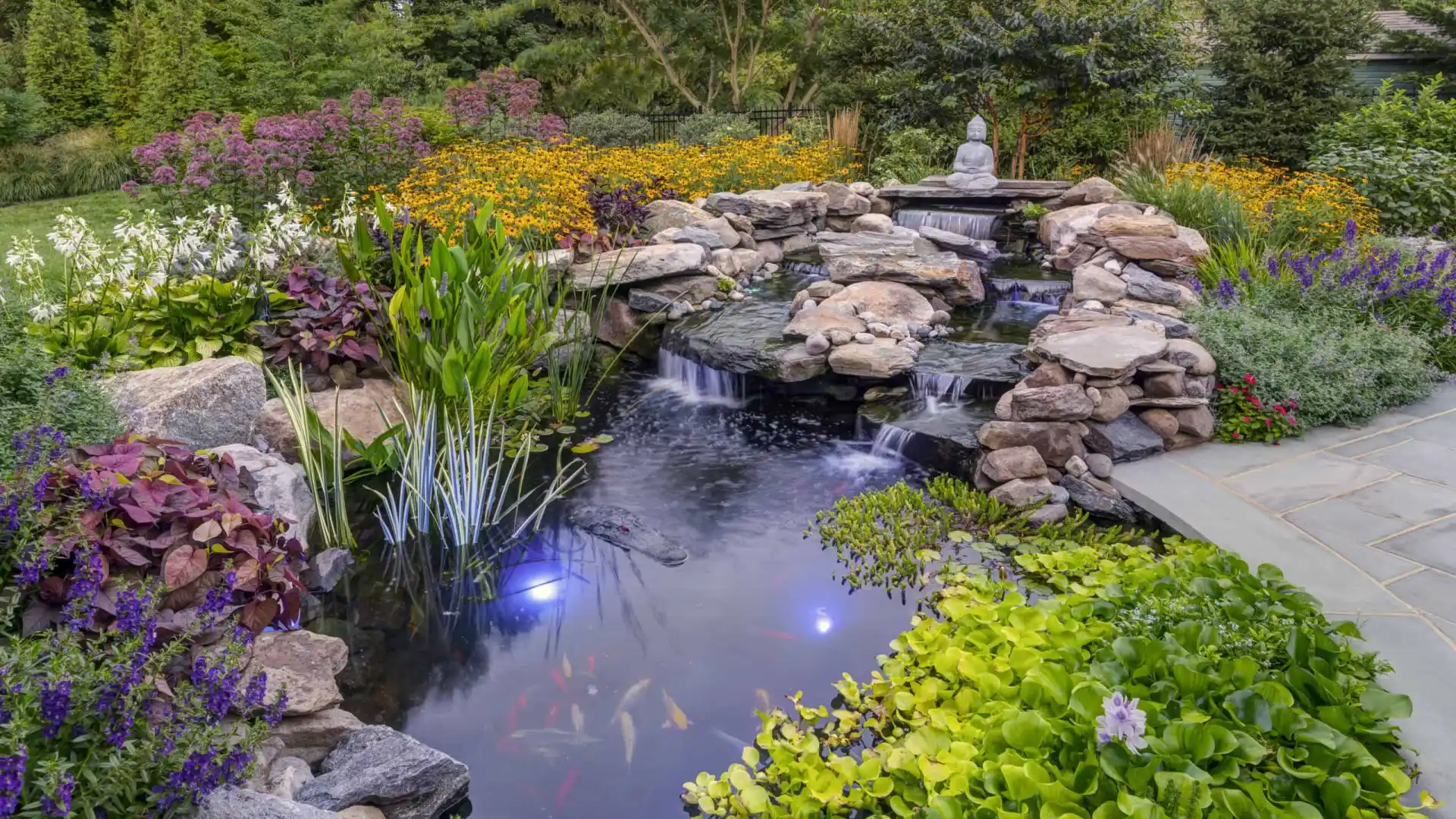 A serene garden scene with a small waterfall flowing into a koi pond surrounded by lush green plants and colorful flowers. A stone figure sits by the water, crafted with hardscaping services from Delaware County PA. Subtle lights illuminate the pond, highlighting the koi fish below.
