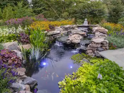 A serene garden scene with a small waterfall flowing into a koi pond surrounded by lush green plants and colorful flowers. A stone figure sits by the water, crafted with hardscaping services from Delaware County PA. Subtle lights illuminate the pond, highlighting the koi fish below.