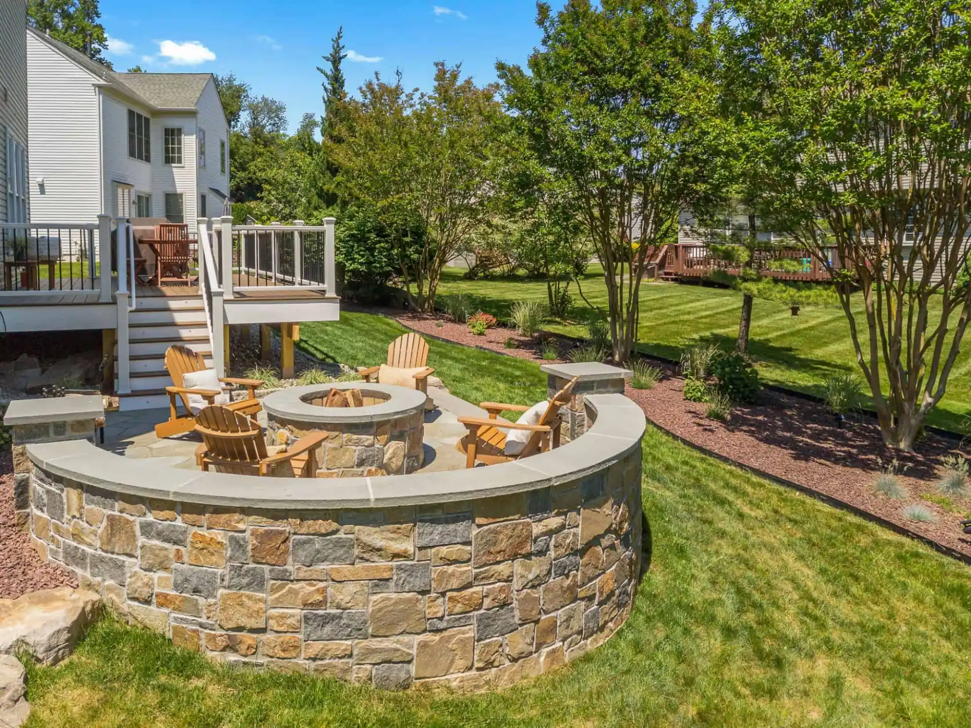 Adirondack chairs encircle a stone fire pit on a patio, enhancing the outdoor living experience in this Delaware County, PA backyard. Bordered by lush grass and pristine landscaping, the scene features a white house with a deck under a clear blue sky.