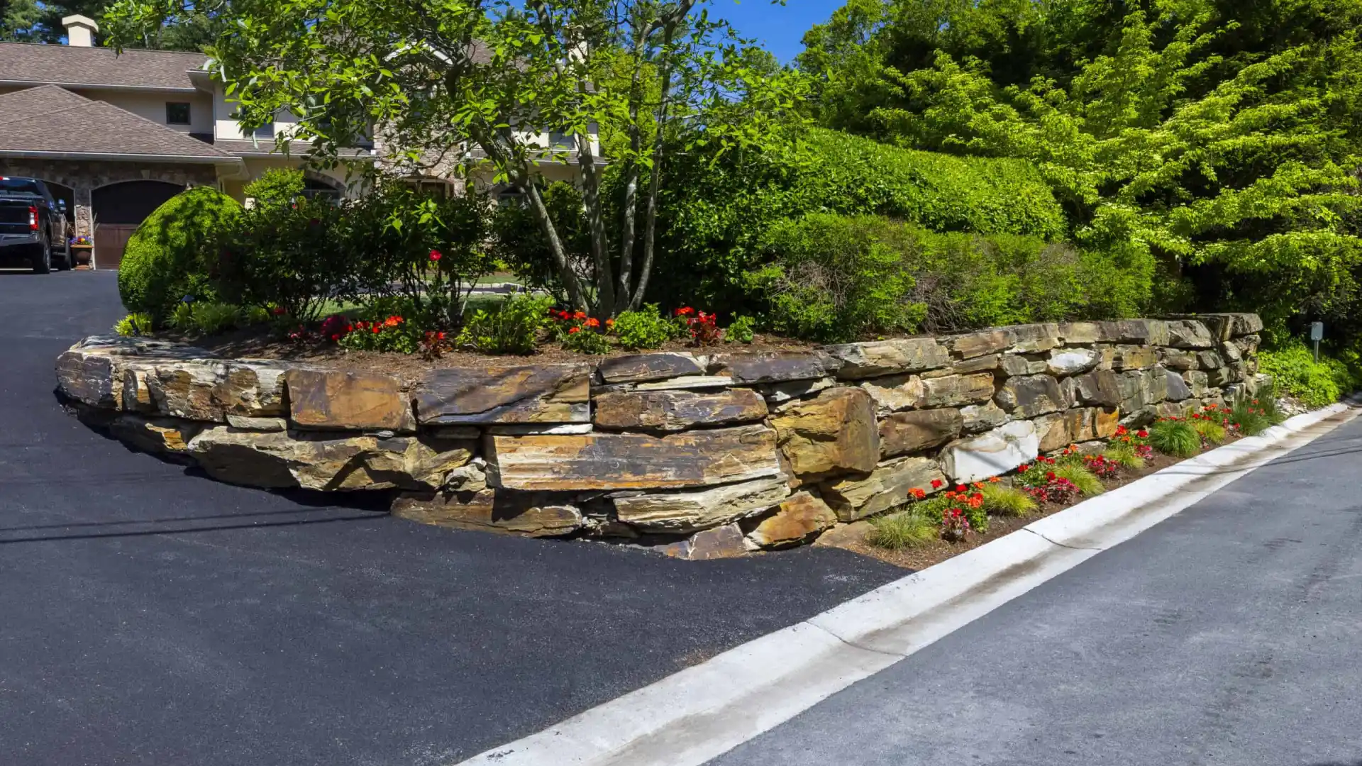 A residential property with a sloped driveway and a stone retaining wall, showcasing expert landscaping services in Delaware County, PA. Lush shrubs and red flowers border the wall. A house with a brown roof is nestled amid vibrant green trees and plants on this bright, sunny day.