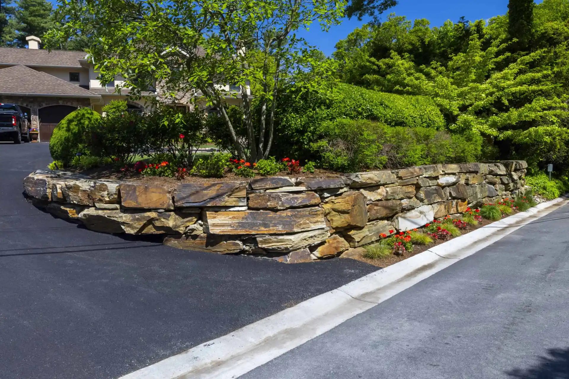 A residential property with a sloped driveway and a stone retaining wall, showcasing expert landscaping services in Delaware County, PA. Lush shrubs and red flowers border the wall. A house with a brown roof is nestled amid vibrant green trees and plants on this bright, sunny day.