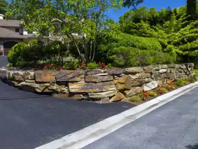 A residential property with a sloped driveway and a stone retaining wall, showcasing expert landscaping services in Delaware County, PA. Lush shrubs and red flowers border the wall. A house with a brown roof is nestled amid vibrant green trees and plants on this bright, sunny day.