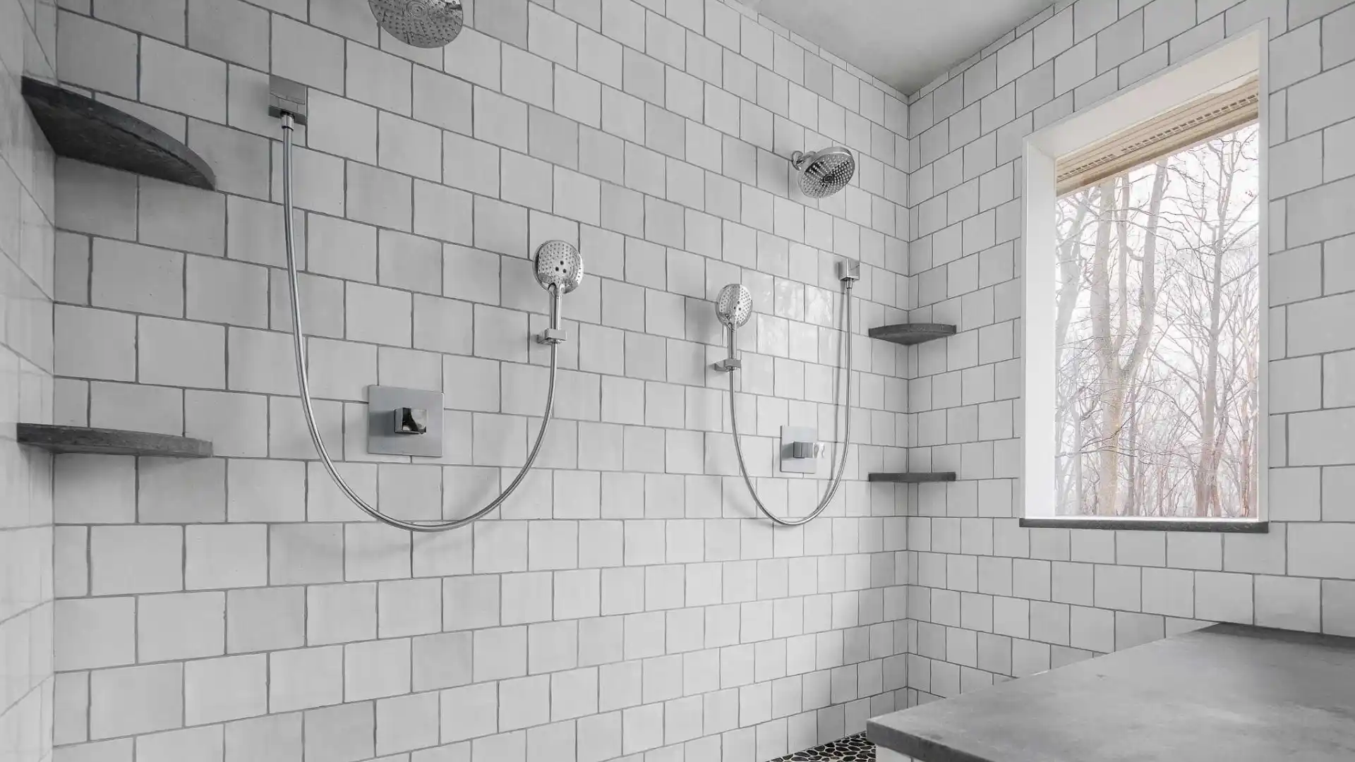 Modern bathroom with two wall-mounted showers over a pebble stone floor. The walls are tiled with white squares, and a window shows bare trees outside. Floating shelves in dark stone complement the design, reminiscent of outdoor living in Delaware County, PA's finest landscapes.