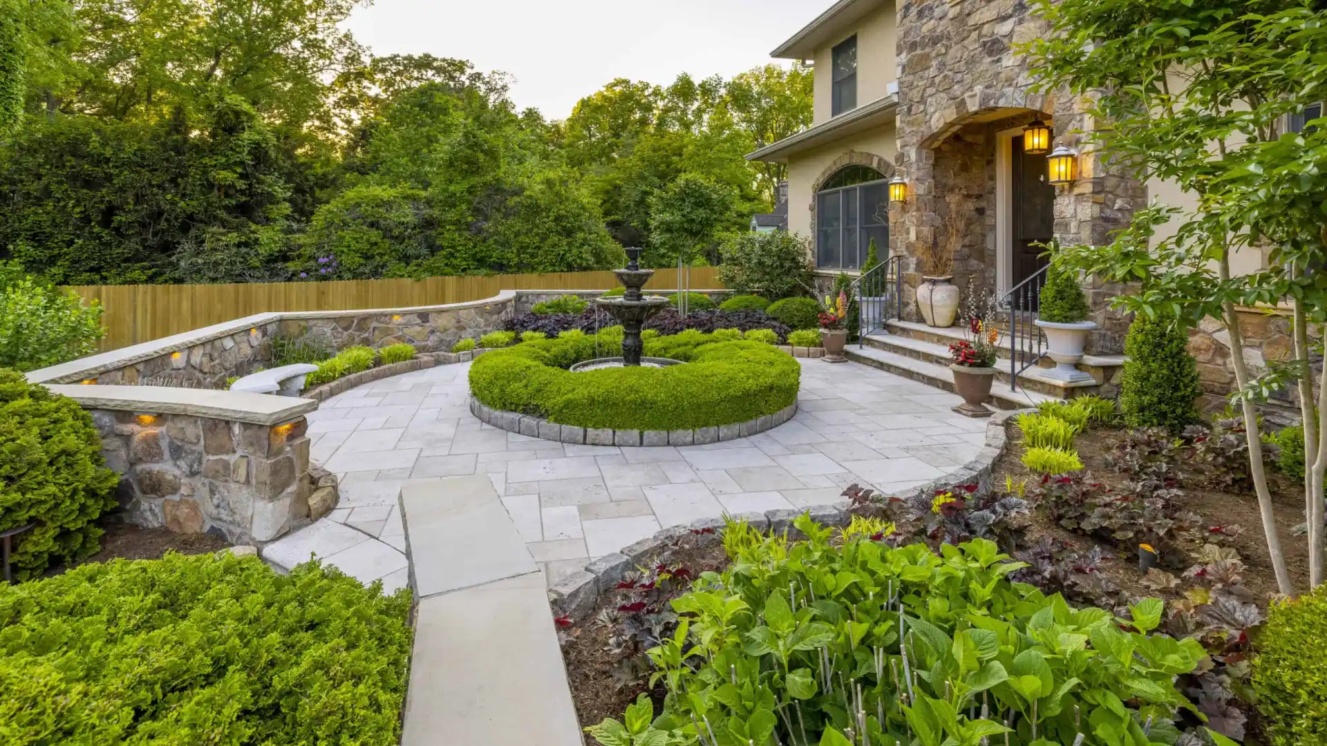 A landscaped garden with a circular stone patio, enhanced by our expert landscaping services in Delaware County, PA, showcases a central fountain. Neatly trimmed hedges and diverse shrubs surround it. A stone house with stairs and flower pots stands amidst lush trees in the background.