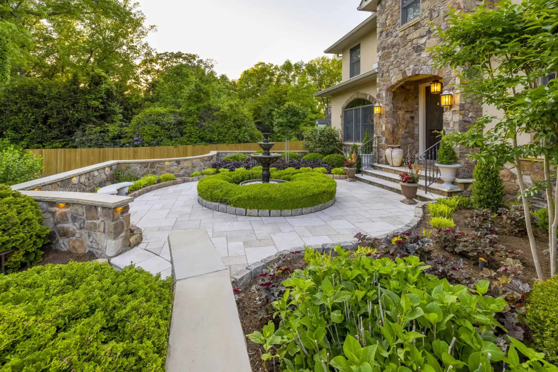 A landscaped garden with a circular stone patio, enhanced by our expert landscaping services in Delaware County, PA, showcases a central fountain. Neatly trimmed hedges and diverse shrubs surround it. A stone house with stairs and flower pots stands amidst lush trees in the background.