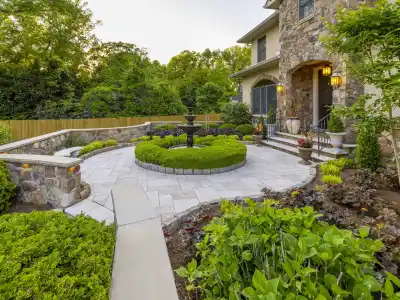 A landscaped garden with a circular stone patio, enhanced by our expert landscaping services in Delaware County, PA, showcases a central fountain. Neatly trimmed hedges and diverse shrubs surround it. A stone house with stairs and flower pots stands amidst lush trees in the background.