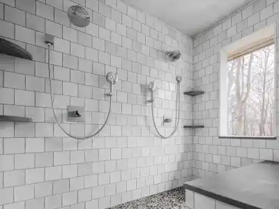 Modern bathroom with two wall-mounted showers over a pebble stone floor. The walls are tiled with white squares, and a window shows bare trees outside. Floating shelves in dark stone complement the design, reminiscent of outdoor living in Delaware County, PA's finest landscapes.