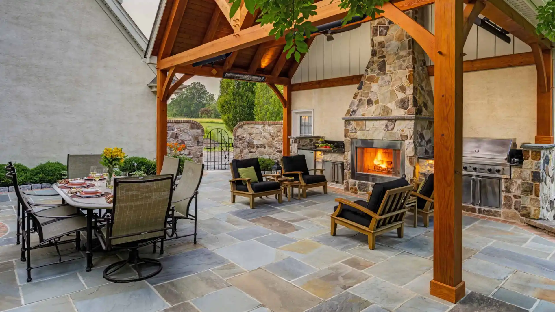 Outdoor living in Delaware County, PA shines with a stone fireplace under a wood pergola. Black-cushioned chairs encircle the fire, while a glass-top dining table invites meals. A built-in grill awaits on the right, and lush greenery envelops this expertly crafted hardscaping haven.