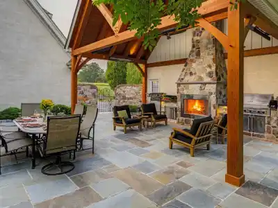 Outdoor living in Delaware County, PA shines with a stone fireplace under a wood pergola. Black-cushioned chairs encircle the fire, while a glass-top dining table invites meals. A built-in grill awaits on the right, and lush greenery envelops this expertly crafted hardscaping haven.