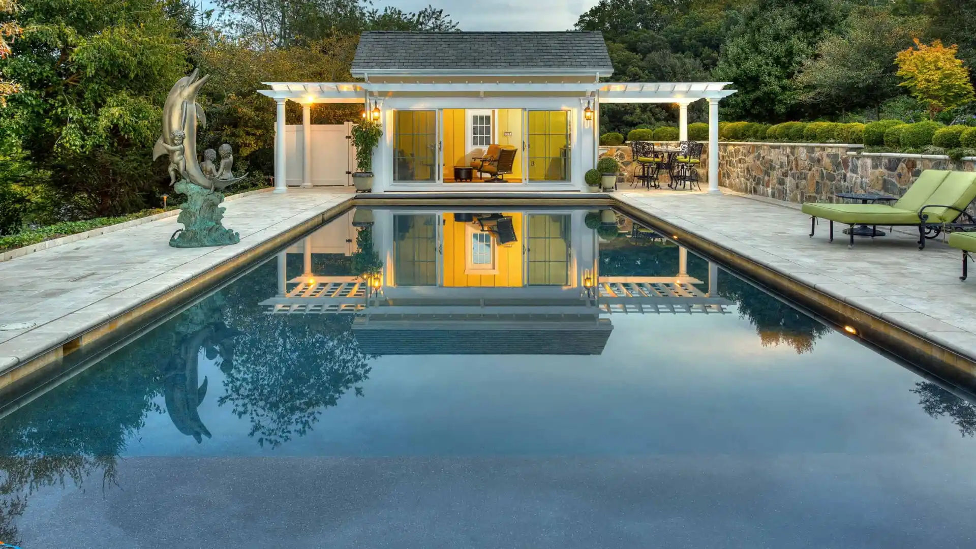 The rectangular pool with clear water reflects a yellow pool house and white pergola. A sculpture is on the left, while trees, bushes, and expertly designed hardscaping services in Delaware County, PA, create a serene escape with lounge chairs on the stone patio to the right.