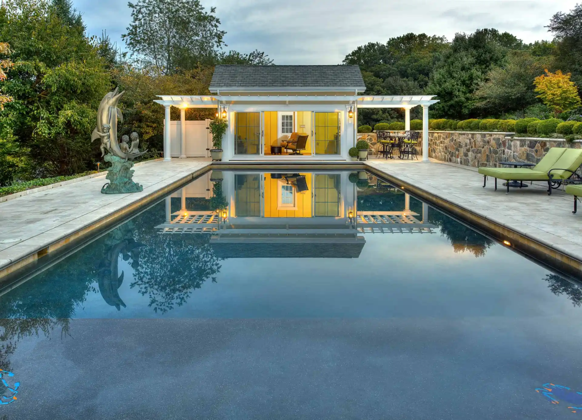 The rectangular pool with clear water reflects a yellow pool house and white pergola. A sculpture is on the left, while trees, bushes, and expertly designed hardscaping services in Delaware County, PA, create a serene escape with lounge chairs on the stone patio to the right.