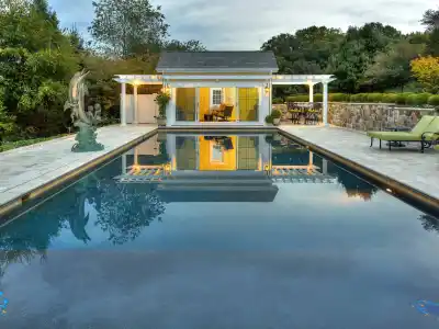 The rectangular pool with clear water reflects a yellow pool house and white pergola. A sculpture is on the left, while trees, bushes, and expertly designed hardscaping services in Delaware County, PA, create a serene escape with lounge chairs on the stone patio to the right.