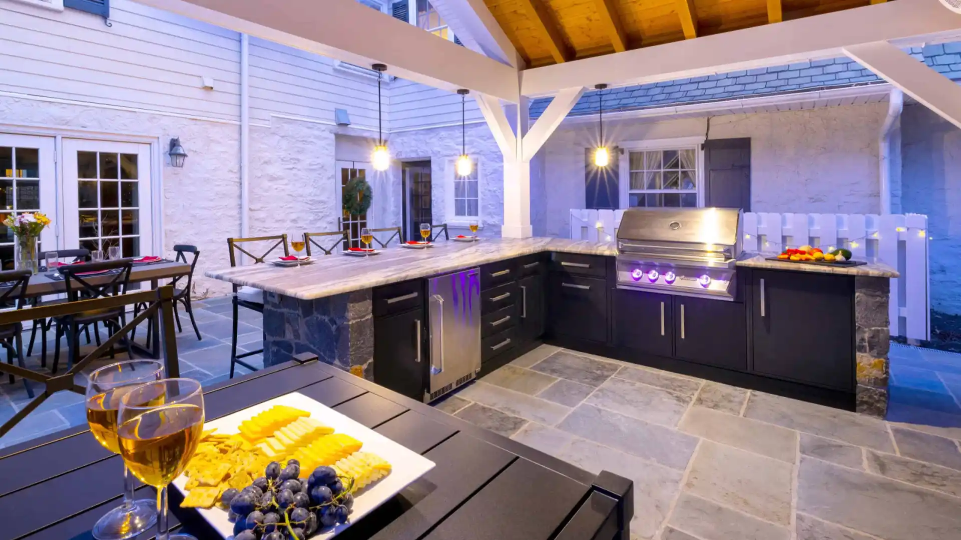 Outdoor living in Delaware County, PA, comes to life with this exquisite setup: a kitchen boasting modern appliances, slate flooring, and wooden ceiling. A bar counter with crackers, cheese, grapes, and wine awaits. A patio dining area is visible amidst refined landscaping services.