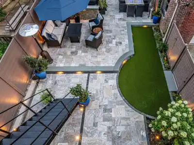 Aerial view of a modern patio in Delaware County, PA, with stone tiles, artificial grass, and cozy seating. Highlighted by a black metal staircase, blue planters, decorative lighting, and a dining set under a dark umbrella. Brick walls wrap around this charming outdoor living space.