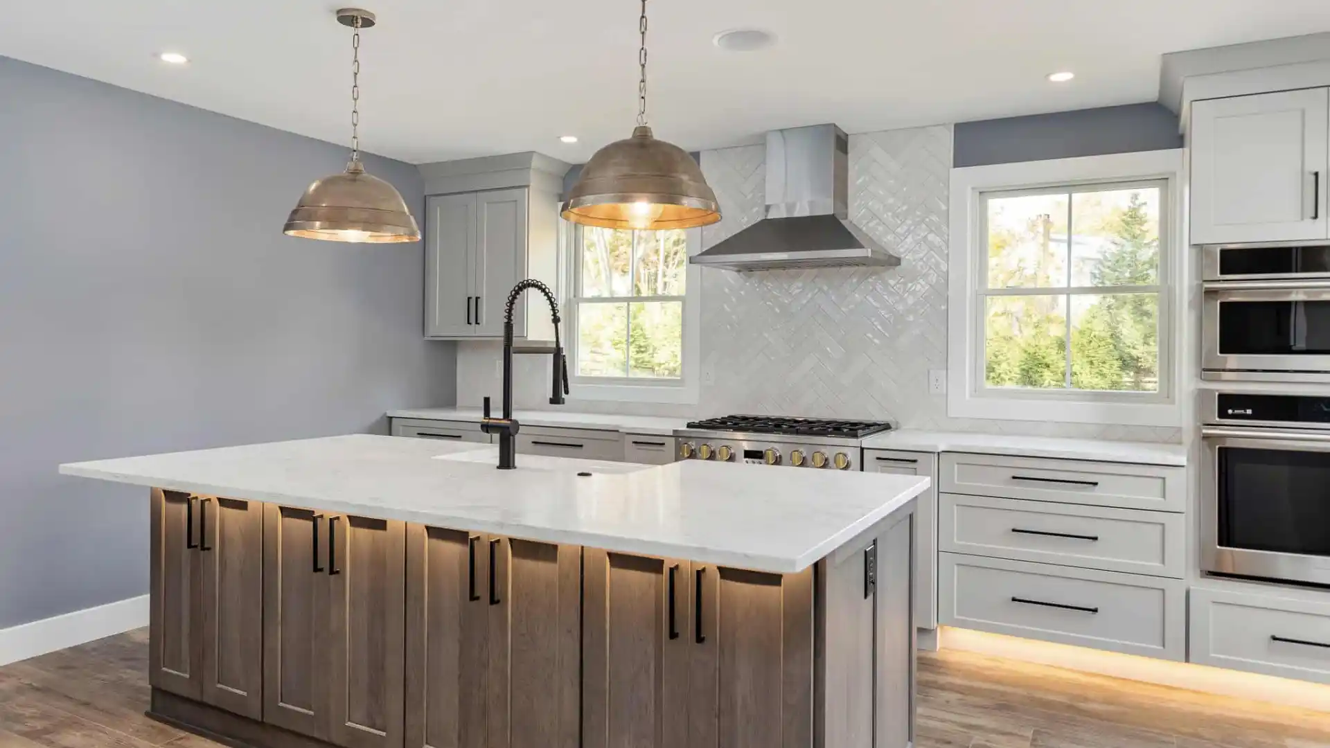 Modern kitchen with a large island featuring dark wood cabinets and a white marble countertop. Two pendant lights hang above. Gray cabinets, stainless steel appliances, and a herringbone backsplash complete the space, blending indoor elegance seamlessly with outdoor living in Delaware County PA.
