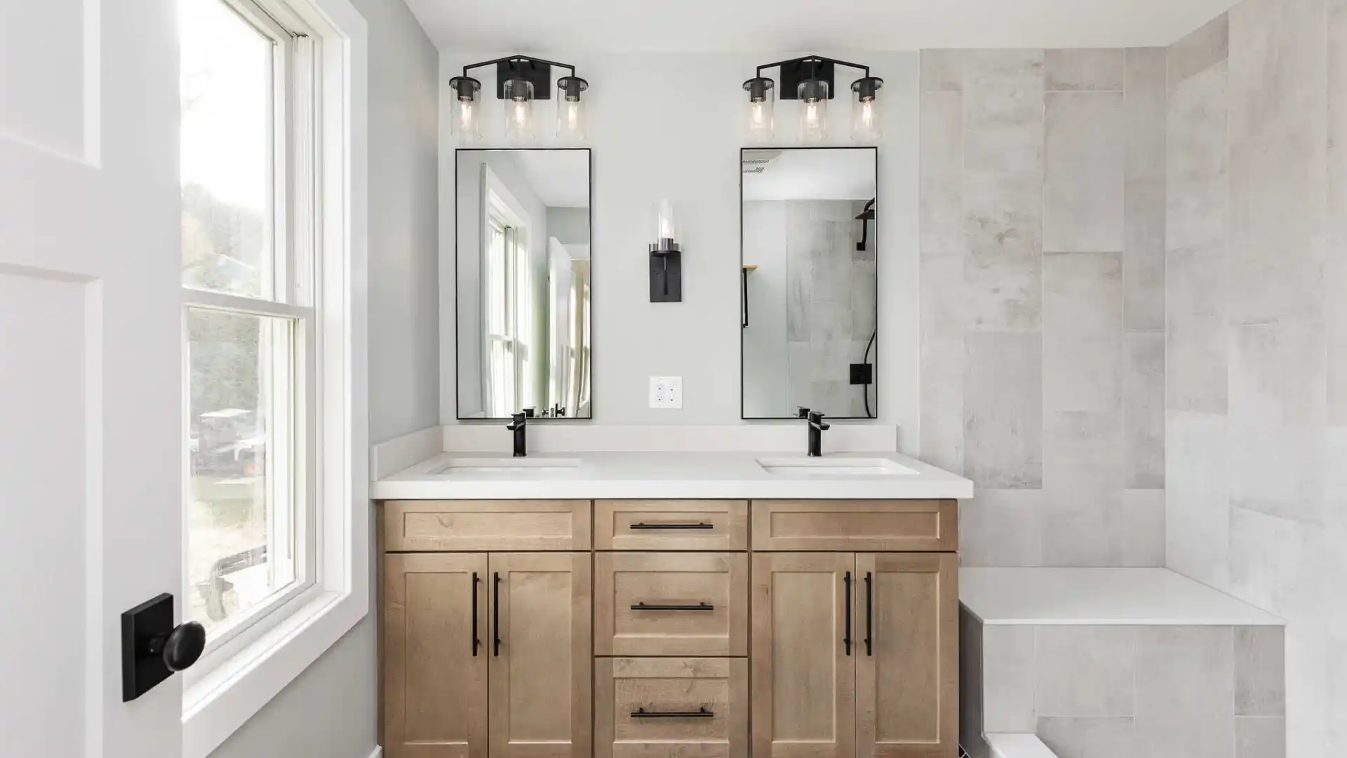 Modern bathroom with light gray walls and a large window on the left. Featuring double sinks with black faucets, light wood cabinetry, and two mirrors, this space exudes tranquility. The dark herringbone tile floor contrasts boldly as if inviting outdoor living sophistication into indoor design.