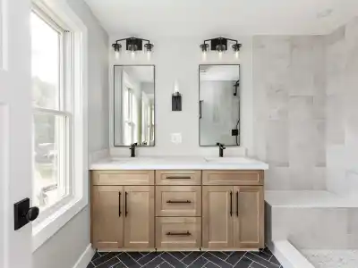 Modern bathroom with light gray walls and a large window on the left. Featuring double sinks with black faucets, light wood cabinetry, and two mirrors, this space exudes tranquility. The dark herringbone tile floor contrasts boldly as if inviting outdoor living sophistication into indoor design.