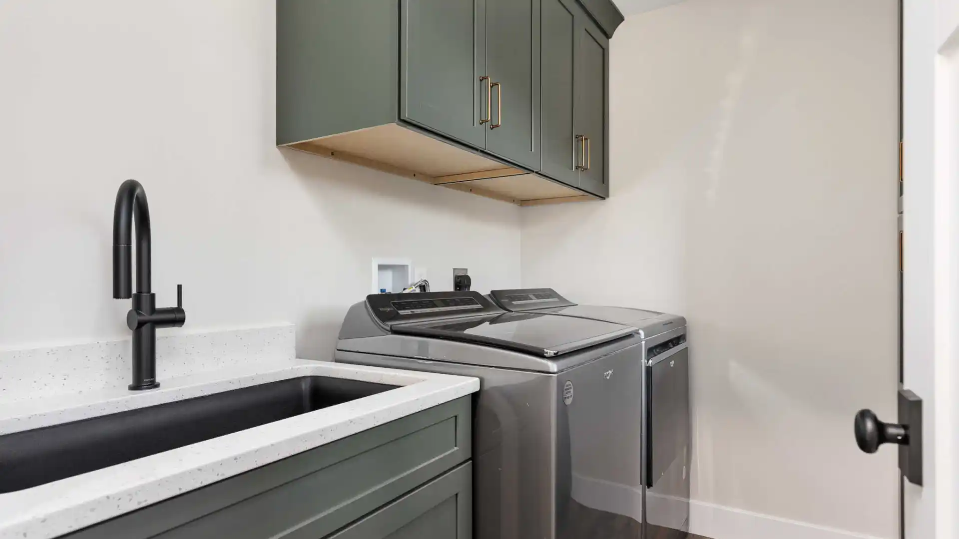Laundry room with dark green cabinets and a countertop holding a gray washing machine and dryer. A black faucet is mounted over a sink on the left. Light walls and wood flooring complete the modern, clean look, reminiscent of outdoor living in Delaware County, PA.