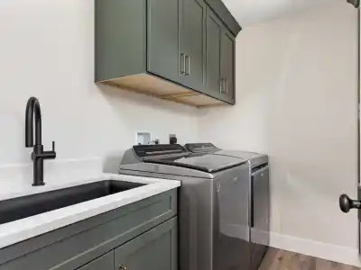 Laundry room with dark green cabinets and a countertop holding a gray washing machine and dryer. A black faucet is mounted over a sink on the left. Light walls and wood flooring complete the modern, clean look, reminiscent of outdoor living in Delaware County, PA.