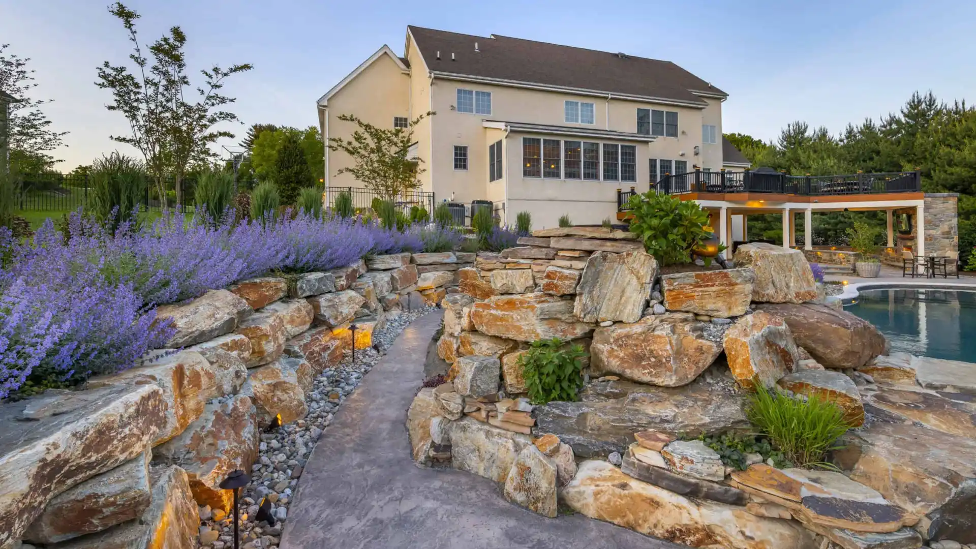 Modern backyard with stone pathway and vibrant landscaping, courtesy of premier landscaping services in Delaware County, PA. Purple flowers and greenery surround a pool and covered outdoor seating area by the house's large windows. Illuminated pathway lights enhance the scene at dusk.