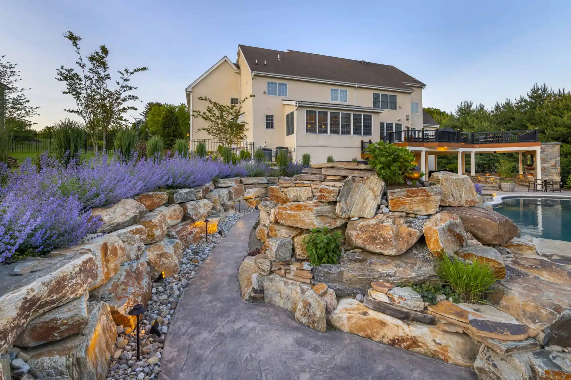 Modern backyard with stone pathway and vibrant landscaping, courtesy of premier landscaping services in Delaware County, PA. Purple flowers and greenery surround a pool and covered outdoor seating area by the house's large windows. Illuminated pathway lights enhance the scene at dusk.