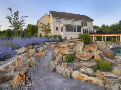Modern backyard with stone pathway and vibrant landscaping, courtesy of premier landscaping services in Delaware County, PA. Purple flowers and greenery surround a pool and covered outdoor seating area by the house's large windows. Illuminated pathway lights enhance the scene at dusk.