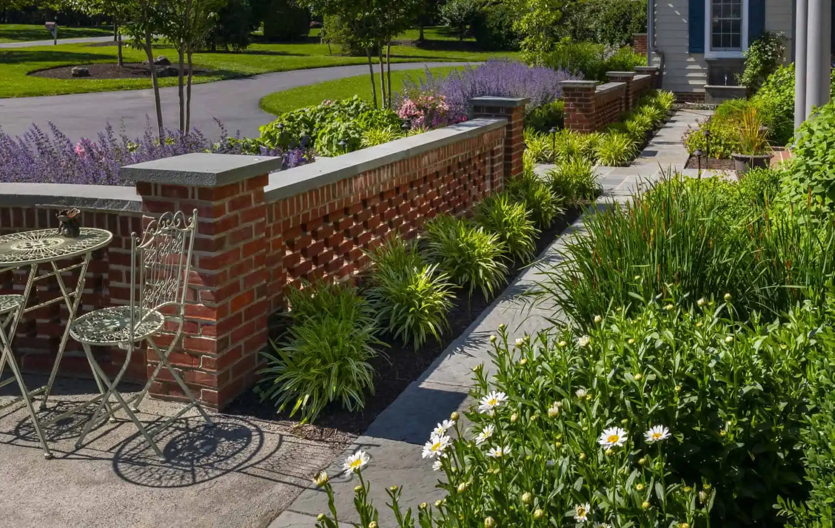 A garden path with lush greenery and blooming white daisies leads to a brick wall. Lavender plants and trimmed bushes line the walkway. An outdoor seating area with metal chairs sits near the path, under a clear blue sky, showcasing expert landscaping services in Delaware County PA.