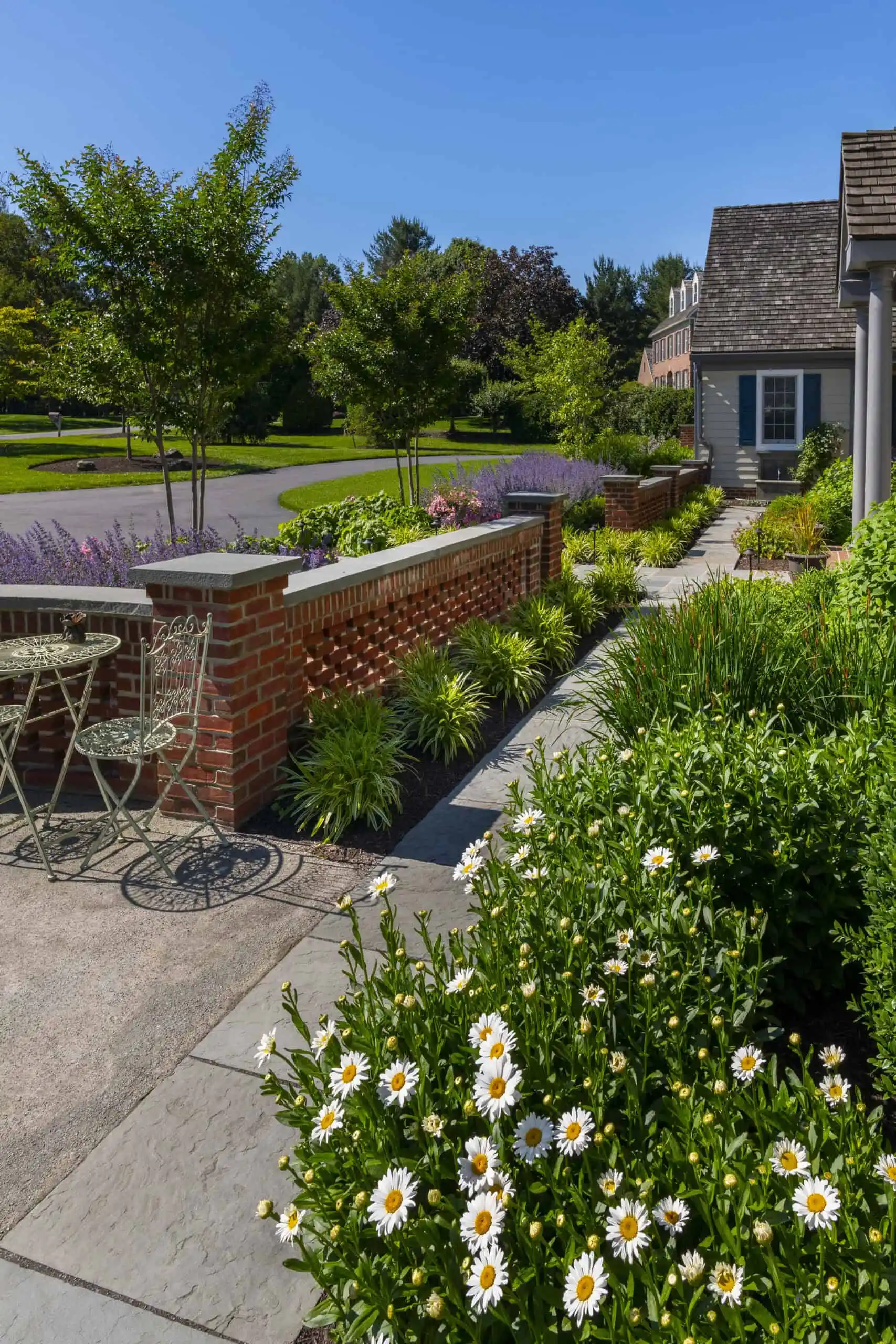 A garden path with lush greenery and blooming white daisies leads to a brick wall. Lavender plants and trimmed bushes line the walkway. An outdoor seating area with metal chairs sits near the path, under a clear blue sky, showcasing expert landscaping services in Delaware County PA.