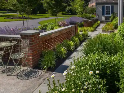 A garden path with lush greenery and blooming white daisies leads to a brick wall. Lavender plants and trimmed bushes line the walkway. An outdoor seating area with metal chairs sits near the path, under a clear blue sky, showcasing expert landscaping services in Delaware County PA.