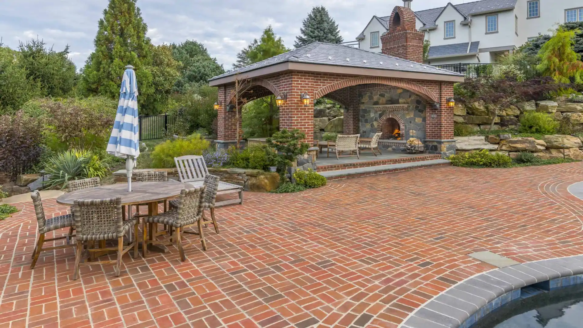 Brick patio featuring a wooden dining set with a blue and white striped umbrella. A covered outdoor fireplace seating area is in the background, enveloped by lush greenery and a stone wall. Experience exquisite outdoor living in Delaware County, PA, with the pool partially visible in the foreground.