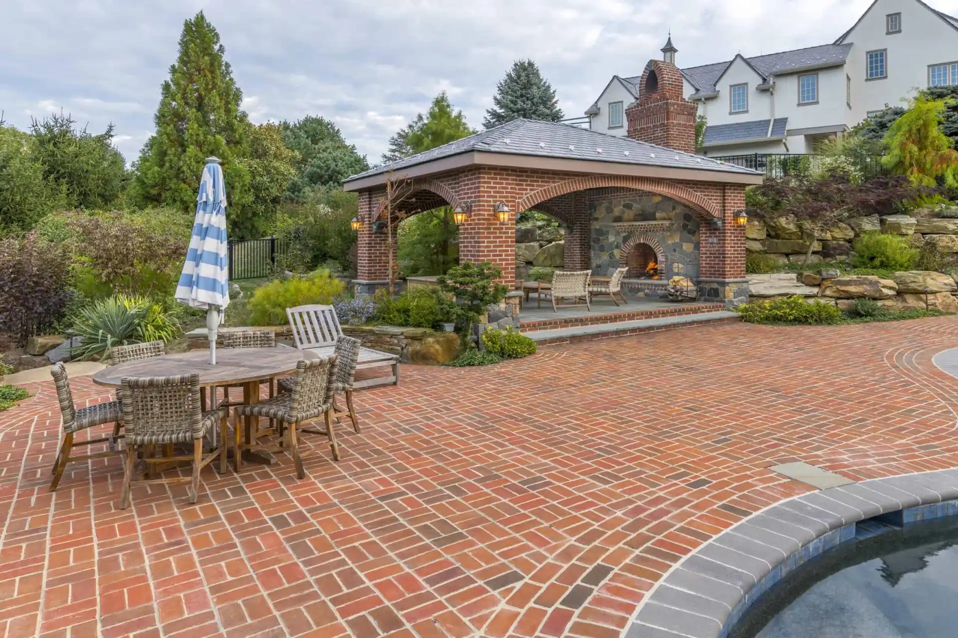 Brick patio featuring a wooden dining set with a blue and white striped umbrella. A covered outdoor fireplace seating area is in the background, enveloped by lush greenery and a stone wall. Experience exquisite outdoor living in Delaware County, PA, with the pool partially visible in the foreground.