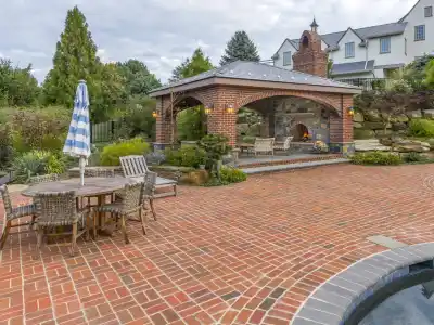 Brick patio featuring a wooden dining set with a blue and white striped umbrella. A covered outdoor fireplace seating area is in the background, enveloped by lush greenery and a stone wall. Experience exquisite outdoor living in Delaware County, PA, with the pool partially visible in the foreground.