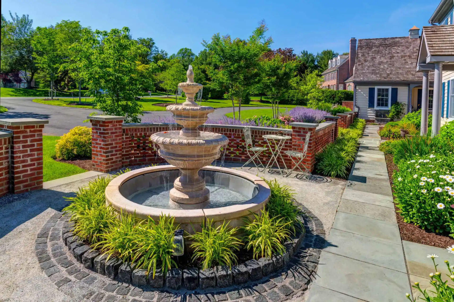 A beautifully landscaped garden, courtesy of premier landscaping services in Delaware County PA, features a stone fountain surrounded by lush greenery. A brick wall encloses the area, where a small patio set invites relaxation. To the right stands a house with a shingled roof under a clear blue sky.