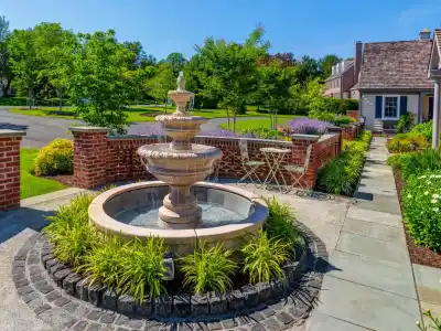 A beautifully landscaped garden, courtesy of premier landscaping services in Delaware County PA, features a stone fountain surrounded by lush greenery. A brick wall encloses the area, where a small patio set invites relaxation. To the right stands a house with a shingled roof under a clear blue sky.