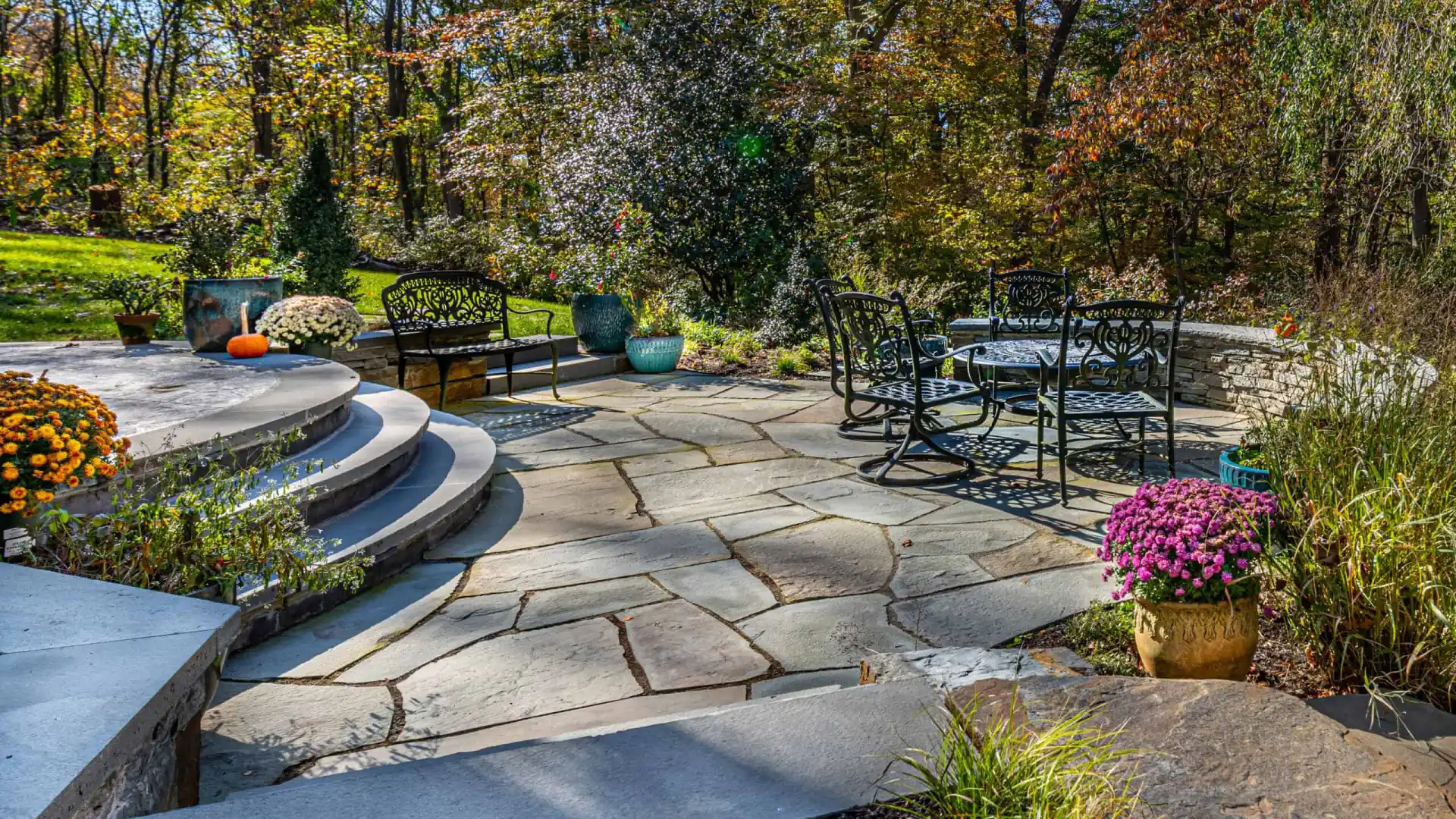 Stone patio with wrought iron furniture, surrounded by colorful potted flowers and greenery. Curved steps lead to a grassy area. Trees with autumn foliage are in the background. Bright sunlight casts shadows across this serene outdoor living space in Delaware County, PA.