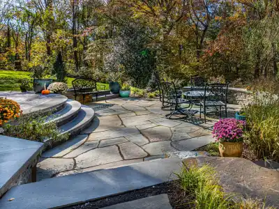 Stone patio with wrought iron furniture, surrounded by colorful potted flowers and greenery. Curved steps lead to a grassy area. Trees with autumn foliage are in the background. Bright sunlight casts shadows across this serene outdoor living space in Delaware County, PA.