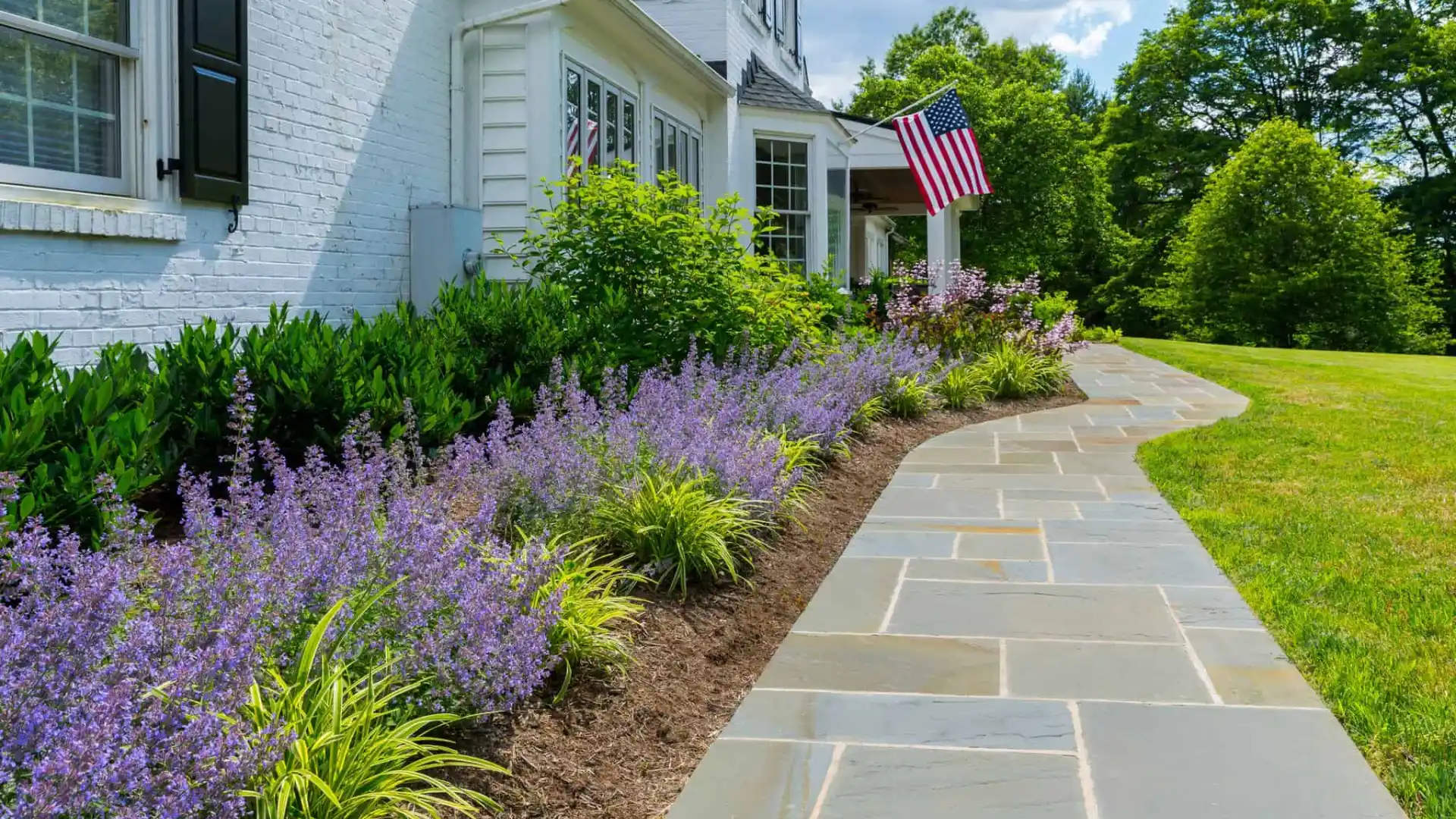 A beautifully landscaped garden beside a white brick house with black shutters showcases outdoor living in Delaware County, PA. Lush greenery and blooming purple flowers line a gray stone pathway. An American flag is displayed near the entrance, with trees standing gracefully in the background.