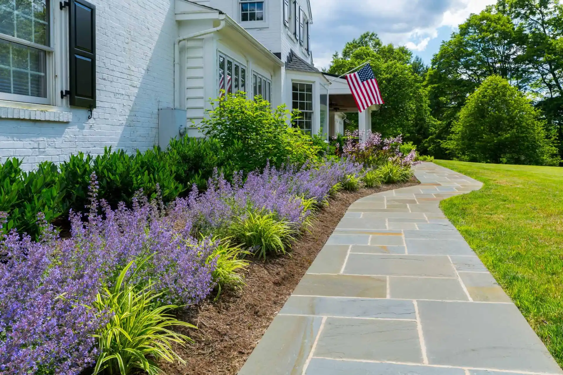 A beautifully landscaped garden beside a white brick house with black shutters showcases outdoor living in Delaware County, PA. Lush greenery and blooming purple flowers line a gray stone pathway. An American flag is displayed near the entrance, with trees standing gracefully in the background.