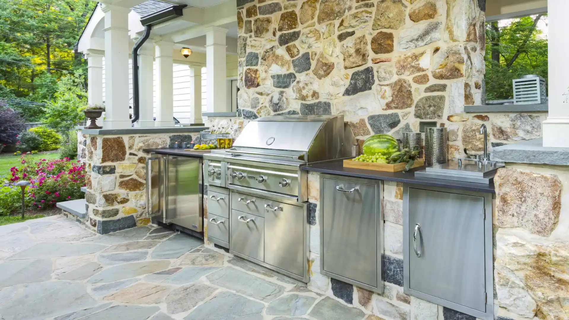 Outdoor kitchen with a stone wall, featuring a stainless steel gas grill, cabinets, and countertop. Nestled within outdoor living in Delaware County, PA, the space is embraced by lush garden blooms and trees, crafting a serene oasis.