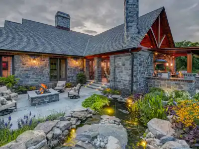 Rustic stone house with a veranda and chimney, warmly lit at dusk. A patio with outdoor seating surrounds a fire pit, perfect for enjoying outdoor living in Delaware County PA. A lush garden and small pond adorned with rocks and greenery decorate the foreground under a cloudy sky.