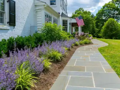A beautifully landscaped garden beside a white brick house with black shutters showcases outdoor living in Delaware County, PA. Lush greenery and blooming purple flowers line a gray stone pathway. An American flag is displayed near the entrance, with trees standing gracefully in the background.