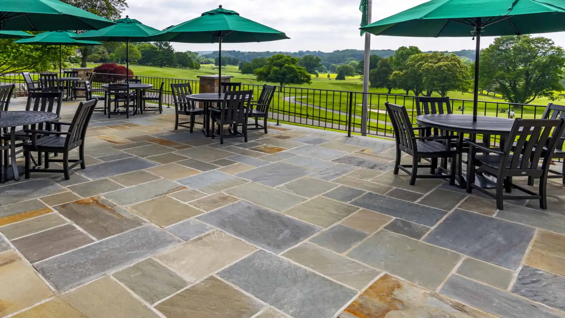 Outdoor patio with stone flooring, a testament to the finesse of hardscaping services in Delaware County, PA. Overlooking lush landscapes, it features tables with dark chairs and large green umbrellas. Trees stretch into a cloudy sky backdrop, embodying serene outdoor living in Delaware County, PA.