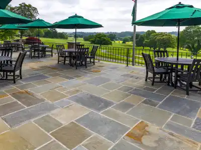Outdoor patio with stone flooring, a testament to the finesse of hardscaping services in Delaware County, PA. Overlooking lush landscapes, it features tables with dark chairs and large green umbrellas. Trees stretch into a cloudy sky backdrop, embodying serene outdoor living in Delaware County, PA.